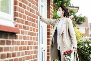 A side view of a female healthcare worker wearing a protective mask knocking on her grandmother's front door, she is dropping off groceries and flowers during the Coronavirus pandemic.